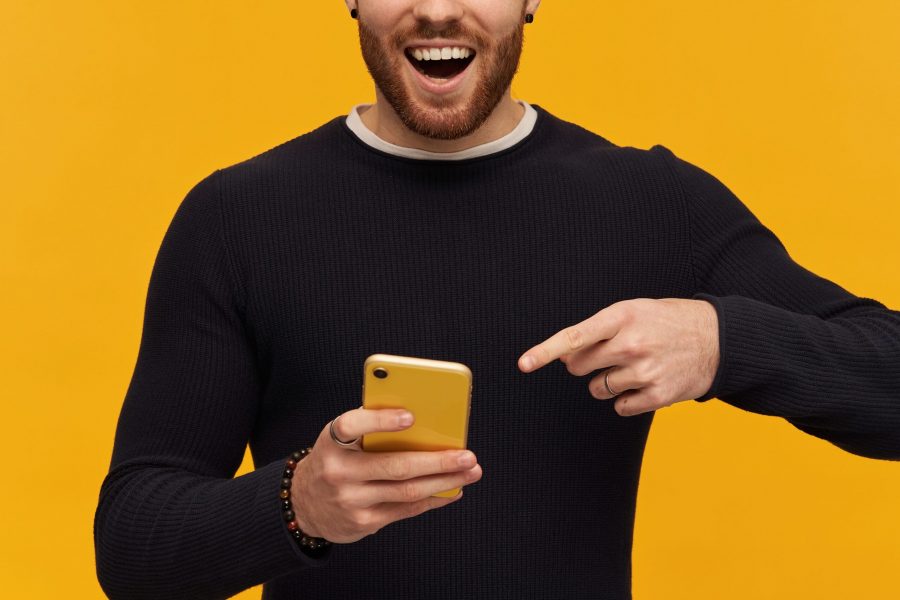 Bearded, happy looking man with brunette hair. Has piercing. Wearing black sweater. Holding and pointing finger at smartphone, copy space. Watching at the camera, isolated over yellow background